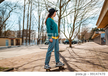 A young Caucasian hipster woman rides down the street on a skateboard. In the background, an alley. Concept of sports lifestyle and street culture A young Caucasian hipster woman rides down the street on a skateboard. In the background, an alley. Concept of sports lifestyle and street culture 99147470