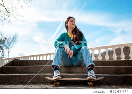 Caucasian teen girl sitting on the steps and leaning her feet on a skateboard. In the background, the blue sky and the Boulevard. Bottom view.Copy space.Concept of sports lifestyle and street culture 99147474