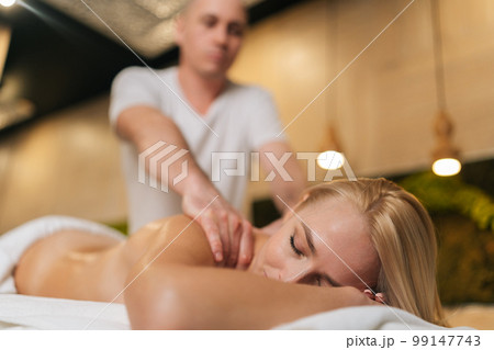 Low-angle view of naked blonde female getting professional relaxing massage at spa salon lying on massage table with closed eyes. Male masseur performing circular movements to relax back muscles. Low-angle view of naked blonde female getting professional relaxing massage at spa salon lying on massage table with closed eyes. Male masseur performing circular movements to relax back muscles. 99147743