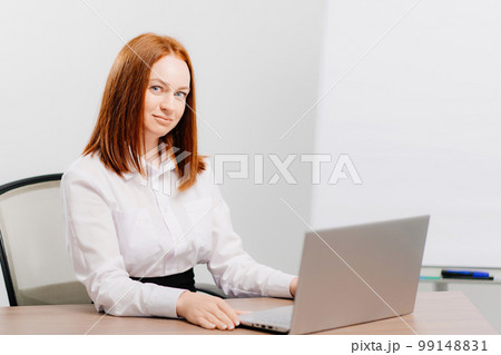 a red-haired woman works at the computer in an office with white walls.  99148831