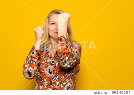 Beautiful blonde woman on vacation in patterned dress on yellow background. Ready to fight with fist defense gesture, confident smiling face, not afraid of trouble. Beautiful blonde woman on vacation in patterned dress on yellow background. Ready to fight with fist defense gesture, confident smiling face, not afraid of trouble. 99149104