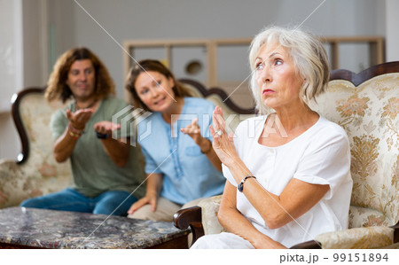 Angry mature female in armchair, keeping distance from the couple sitting on the couch at home Angry mature female in armchair, keeping distance from the couple sitting on the couch at home 99151894