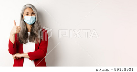 Coronavirus and business concept. Asian female entrepreneur in face mask looking cheerful, showing thumb-up in approval, praising using protective measures at work Coronavirus and business concept. Asian female entrepreneur in face mask looking cheerful, showing thumb-up in approval, praising using protective measures at work 99158901