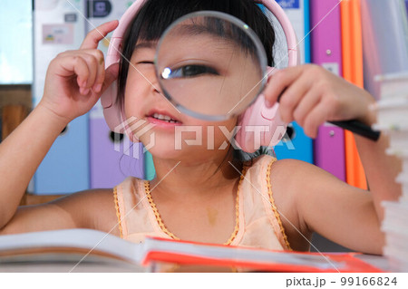 Cheerful little girl in headphones reading a book with a magnifying glass sitting at the table in her room at home. 99166824