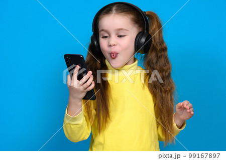 Portrait of a little girl with a phone and wireless headphones, a child in a yellow sweater on a blue background. 99167897