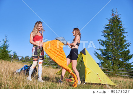 Two women hikers camping outdoors. Female tourists inflating the air mattress, wearing sportswear, hiking in mountains, making campsite. Concept of tourism and traveling. 99173527