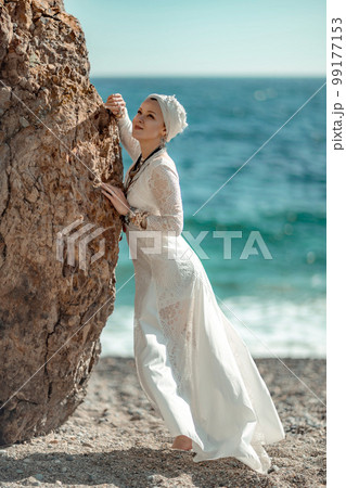 Woman white dress sea stones rocks.Middle-aged woman looks good with blond hair, boho style in a white long dress on beach jewelry around her neck and arms. 99177153