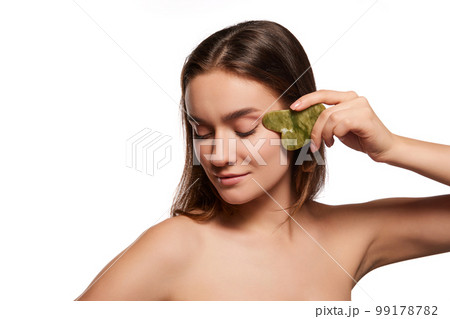 Young beautiful girl massaging face with Gua sha over white studio background. Circulation and lifting effect. Toned and smooth skin  99178782