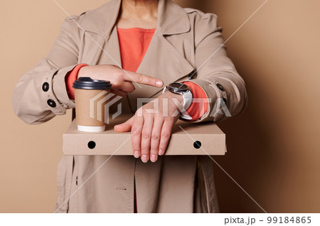 It's lunch time. Close-up woman holding takeaway pizza and coffee in paper mug, points her index finger at a wristwatch 99184865