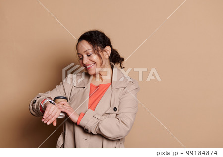 Beautiful woman checking time on her wristwatch, happy to be punctual for a meeting, isolated on beige background Beautiful woman checking time on her wristwatch, happy to be punctual for a meeting, isolated on beige background 99184874