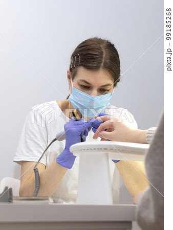 White nail technician in mask drills, removes client's cuticle with hardware machine, holding nail brush. Customer wears warm cloth, sweater. Combined manicure, beauty treatment in spa. Vertical White nail technician in mask drills, removes client's cuticle with hardware machine, holding nail brush. Customer wears warm cloth, sweater. Combined manicure, beauty treatment in spa. Vertical 99185268