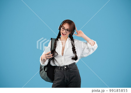 Happy caucasian teenager girl with pigtails in glasses with headphones student with backpack uses phone Happy caucasian teenager girl with pigtails in glasses with headphones student with backpack uses phone 99185855