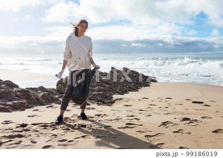 World environment day. Female volunteer standing on beach with bag, picking up garbage and cleaning the coastal zone World environment day. Female volunteer standing on beach with bag, picking up garbage and cleaning the coastal zone 99186019