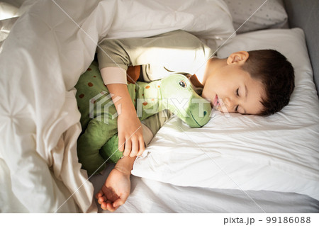 Tired european small kid in pajamas sleeps on bed with dinosaur toy in bedroom, top view, empty space Tired european small kid in pajamas sleeps on bed with dinosaur toy in bedroom, top view, empty space 99186088