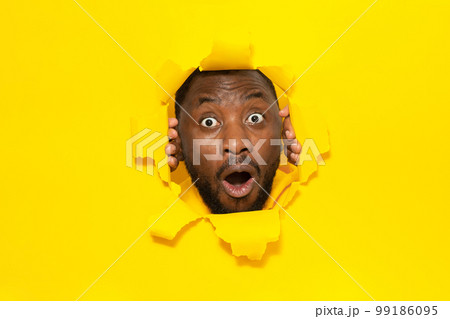 Omg. Shocked black man looking at camera with open mouth through hole in torn yellow paper background 99186095