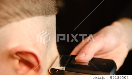 close up men's hairstyling and haircutting with barber shaving machine, hair cut for male. Hairdresser service with shaver in a modern barbershop in a dark key lightning with warm light side view close up men's hairstyling and haircutting with barber shaving machine, hair cut for male. Hairdresser service with shaver in a modern barbershop in a dark key lightning with warm light side view 99186502