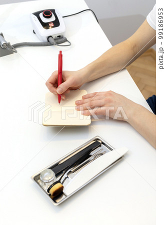 Unrecognizable nail master makes notes in notebook with pen, sitting at workstation, table in nail beauty treatment salon. Professional tools are on white table. Manicurist's routine. Vertical Unrecognizable nail master makes notes in notebook with pen, sitting at workstation, table in nail beauty treatment salon. Professional tools are on white table. Manicurist's routine. Vertical 99186953
