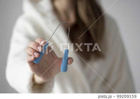 Metal tongue scraper tool in hand against the background of a woman in a bathrobe. Metal tongue scraper tool in hand against the background of a woman in a bathrobe. 99209159