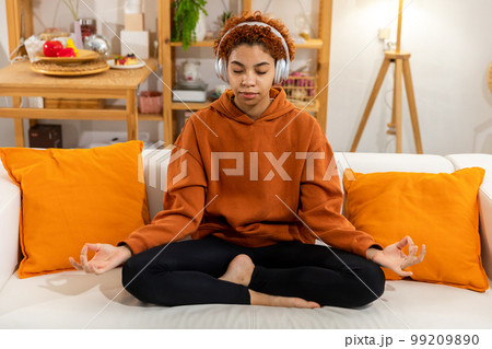 Yoga mindfulness meditation. Young healthy african girl practicing yoga at home. Woman sitting in lotus pose on couch meditating smiling relaxing indoor. Girl doing breathing practice. Yoga at home 99209890