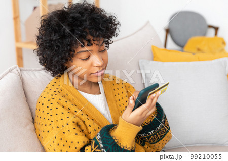 African american woman shopping online holding smartphone paying with gold credit card. Girl sitting at home buying on Internet enter credit card details. Online shopping ecommerce delivery service African american woman shopping online holding smartphone paying with gold credit card. Girl sitting at home buying on Internet enter credit card details. Online shopping ecommerce delivery service 99210055