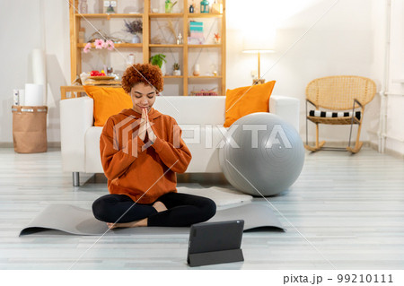 Yoga mindfulness meditation. Young healthy african girl practicing yoga at home. Woman sitting in lotus pose on yoga mat eyes close hands in prayer meditating indoor. Girl doing breathing practice 99210111