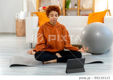 Yoga mindfulness meditation. Young healthy african girl practicing yoga at home. Woman sitting in lotus pose on yoga mat meditating smiling relaxing indoor. Girl doing breathing practice. Yoga at home 99210115