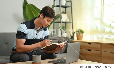 Millennial man using laptop nd making important notes on notepad, working online from home Millennial man using laptop nd making important notes on notepad, working online from home 99211075