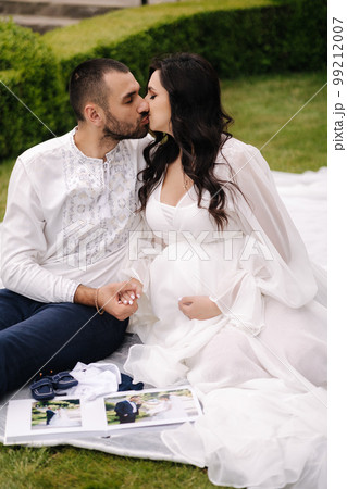 Portrait of beautiful couple sitting on a plaid on the grass. Elegant pregnant woman with husband waiting for baby. Future parents 99212007