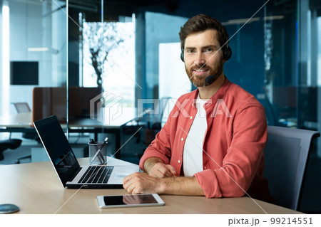 Portrait of mature businessman in red shirt inside office, man with beard and video call headset smiling and looking at camera, online customer support worker with laptop. Portrait of mature businessman in red shirt inside office, man with beard and video call headset smiling and looking at camera, online customer support worker with laptop. 99214551