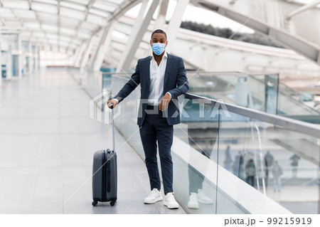 Black Man Wearing Suit And Medical Mask Posing With Suitcase At Airport Black Man Wearing Suit And Medical Mask Posing With Suitcase At Airport 99215919