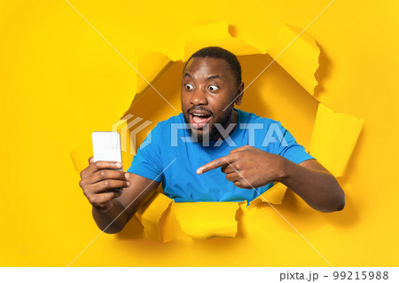 Shocked black man holding smartphone and pointing on it, looking at screen, standing through yellow paper torn hole Shocked black man holding smartphone and pointing on it, looking at screen, standing through yellow paper torn hole 99215988
