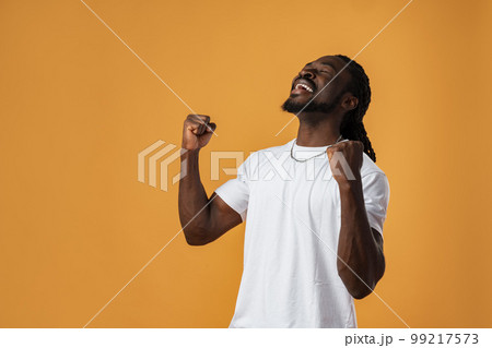 Portrait of excited african guy celebrating success with clenched fists against yellow background Portrait of excited african guy celebrating success with clenched fists against yellow background 99217573