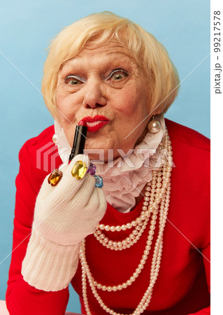Applying lipstick. Beautiful old woman, grandmother in stylish red dress and pearl necklace posing over blue studio background. Concept of age, fashion and emotions Applying lipstick. Beautiful old woman, grandmother in stylish red dress and pearl necklace posing over blue studio background. Concept of age, fashion and emotions 99217578