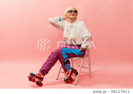 Retro style. Beautiful old woman, grandmother in stylish sportive trousers posing on vintage rollers over pink studio background. Concept of age, fashion and emotions 99217621
