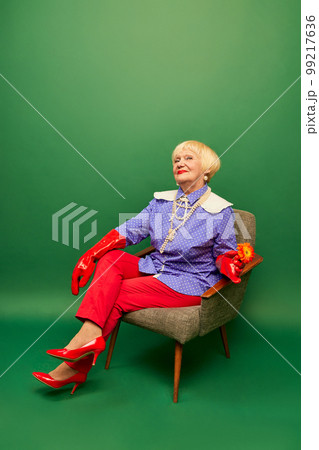 Red and purple. Beautiful old woman, grandmother in stylish colorful clothes sitting on armchair over green studio background. Concept of age, fashion and emotions Red and purple. Beautiful old woman, grandmother in stylish colorful clothes sitting on armchair over green studio background. Concept of age, fashion and emotions 99217636