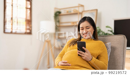 Happy Asian pregnant woman using her phone while relaxing on sofa in her living room Happy Asian pregnant woman using her phone while relaxing on sofa in her living room 99219572