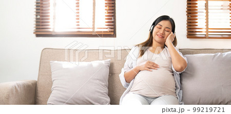 Happy Asian pregnant woman listening to music through her headphones on sofa. pregnancy lifestyle 99219721
