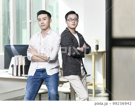 portrait of two young asian business men posing back to back in office looking at camera portrait of two young asian business men posing back to back in office looking at camera 99236942