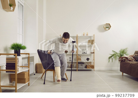 Man with a broken leg tries to stand up off his chair and walk with a stick and crutches 99237709