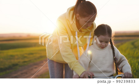 mom teaches ittle child ride bike summer road park sunset. happy family concept. cheerful child pedals bicycle sun. kid controls steering wheel two-wheeled bicycle summer sunset. weekends motherhood mom teaches ittle child ride bike summer road park sunset. happy family concept. cheerful child pedals bicycle sun. kid controls steering wheel two-wheeled bicycle summer sunset. weekends motherhood 99239833