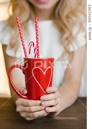 A red mug with a heart pattern with cocoa and paper tubes in the hands of a woman. Valentine's Day Concept 99241463