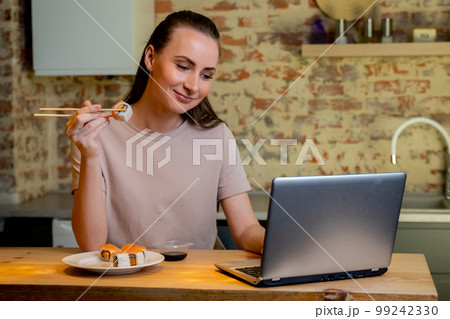Young pretty woman sitting at her kitchen eat sushi at table home working with a laptop 99242330