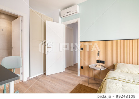 Bedroom with bed, wooden wardrobe built into wall, table and chair for work. Air conditioning on door for comfortable temperature in room. Near bed there is a table with night lamp. 99243109