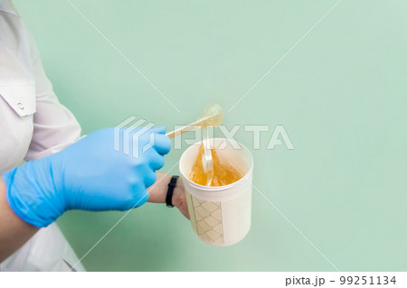 The cosmetologist holds a jar of sugar wax, shows texture of paste with a spatula The cosmetologist holds a jar of sugar wax, shows texture of paste with a spatula 99251134