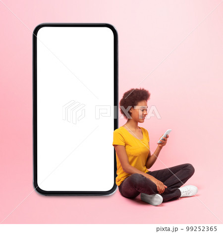 Smiling young black curly lady sit on floor with phone near big smartphone with empty screen 99252365