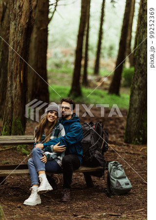 Glad millennial caucasian husband in jacket hugging wife, resting on bench in cold forest with backpack 99252369