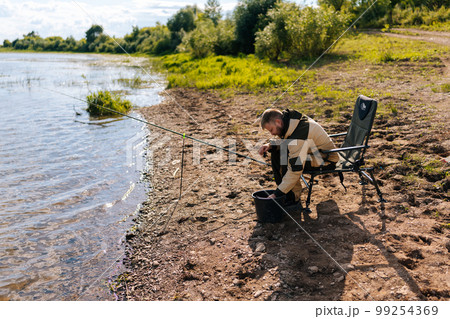 Side view of bearded fisherman wearing raincoat preparing fishing gear on hook sitting on travel chair on river bank alone. Concept of lifestyle, leisure activity on nature. 99254369