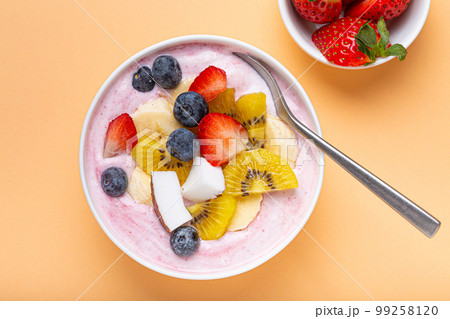 Healthy breakfast or dessert yogurt bowl with fresh banana, strawberry, blueberry, cocos, kiwi top view on minimal pastel paper background and spoon 99258120