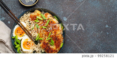 Asian noodles ramen soup, deep fried panko chicken fillet and boiled eggs in ceramic bowl with chop sticks and soy sauce on stone rustic background top view, space for text 99258121