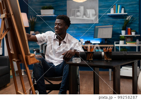 Smiling african american male cartoonist using colored pencils to sketch on easel at art studio. Wheelchair user drawing happily on canvas in bedroom. Person with artistic skills working in atelier. 99258287